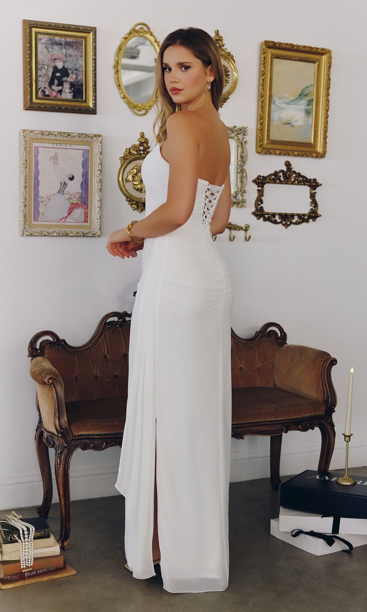 A woman wearing the Amoris T726W, a long off-white formal dress with an adjustable lace-up back, stands beside a vintage-style couch. Framed artwork and mirrors add to the elegant ambiance behind her.